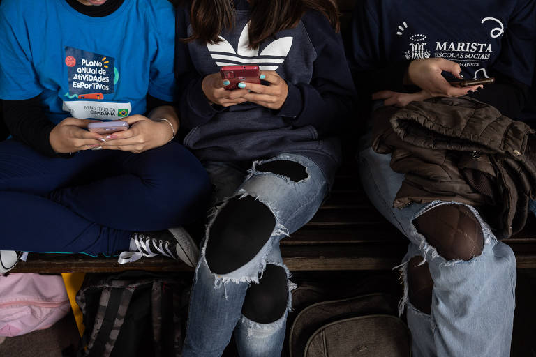 Foto mostra três adolescentes manuseando aparelhos celulares enquanto estão sentadas em um banco. A foto não mostra o rosto das estudantes