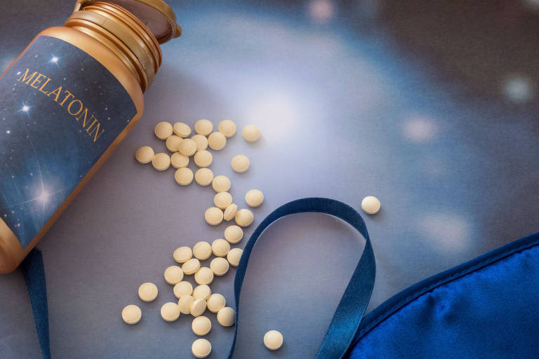 Fotografia colorida mostra um frasco escrito "Melatonin", com vários comprimidos brancos para fora; eles estão sobre um lençol azul, ao lado de uma máscara de dormir da mesma cor