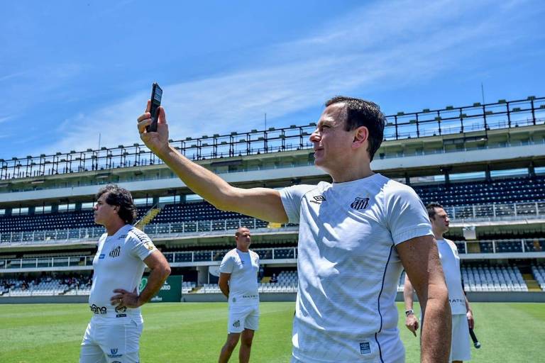 João Doria está tirando uma selfie no campo de um estádio de futebol. Ele veste o uniforme do Santos. Ao redor deles, há outras pessoas vestidas com o uniforme do time.