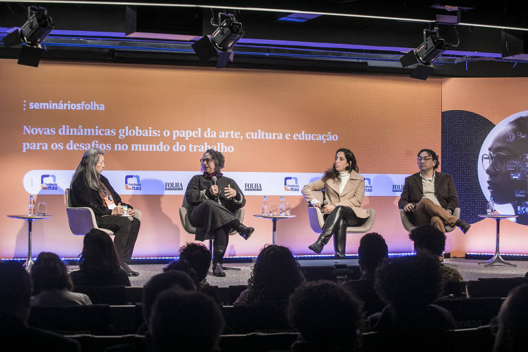 Três mulheres e um homem estão sentados em cadeiras dispostas no palco de um auditório; o fundo é laranja e contém o nome do seminário: 'Novas dinâmicas globais: o papel da arte, cultura e educação para os desafios no mundo do trabalho'