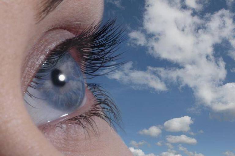 Fotografia colorida foca os olhos azuis de uma mulher branca; ao fundo há um céu no mesmo tom de azul e nuvens brancas