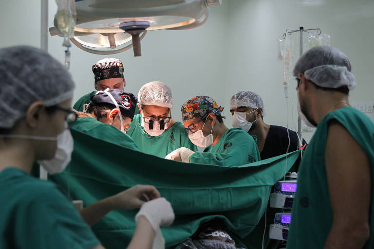 Seis médicos durante procedimento cirúrgico de transplante de fígado. Não é possível ver o procedimento, há uma manta verde na frente. 