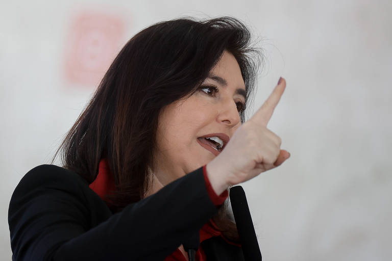 A ministra do Planejamento, Simone Tebet, em Brasília; ela é uma mulher branca, de cabelos lisos e escuros, veste uma jaqueta preta, camisa vermelha e está com o dedo indicador direito levantado