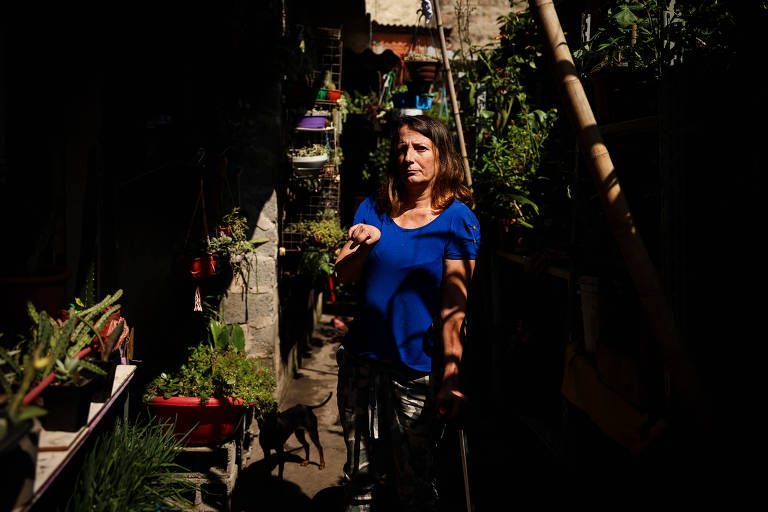 Mulher de 53 anos, com camiseta azul, está no centro da área externa de uma casa, cercada por plantas