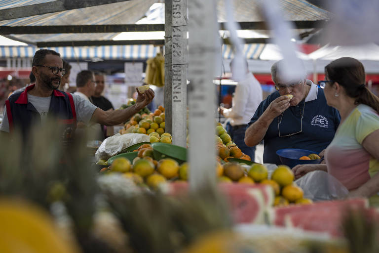 Imagem mostra compradores em uma feira de frutas. Eles estão em uma volta de uma banca cheia de laranjas