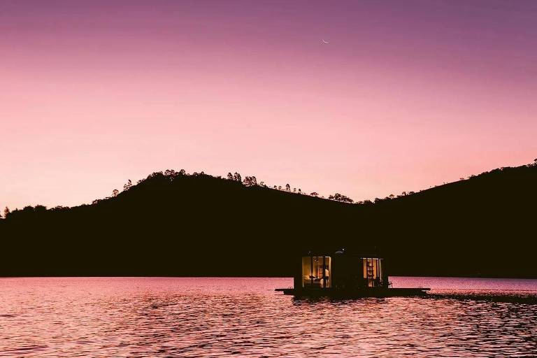 Fotografia colorida mostra uma pequena casa em um lago com montanhas ao fundo durante um por-do-sol cor-de-rosa