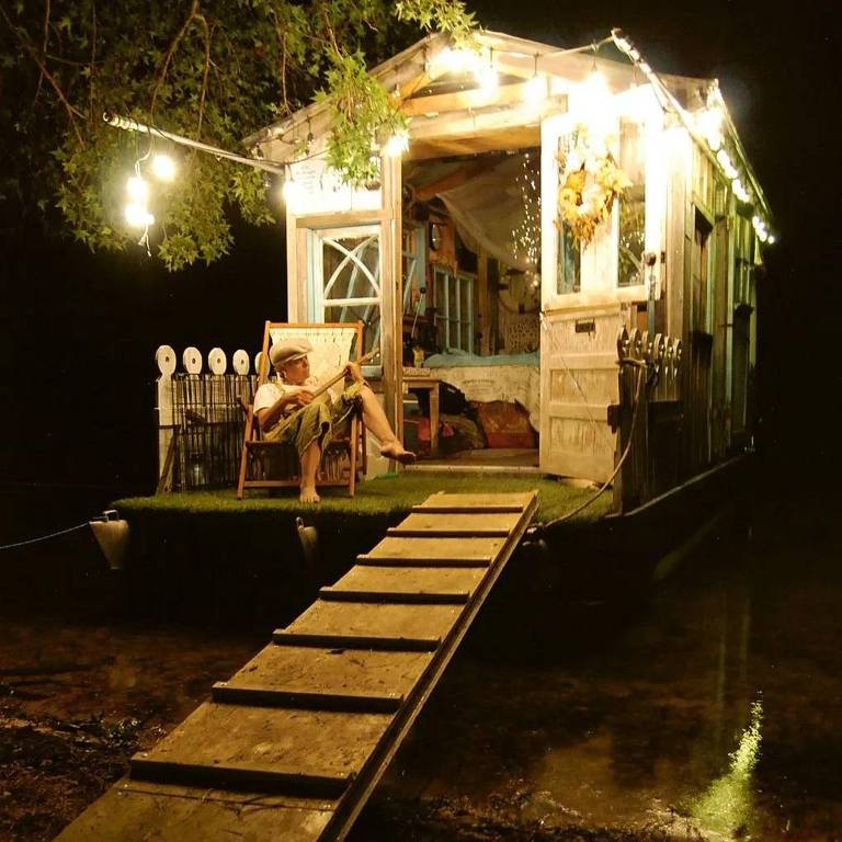 Foto mostra cabana em rio à noite iluminada por luzinhas e com uma mulher tocando banjo na varanda