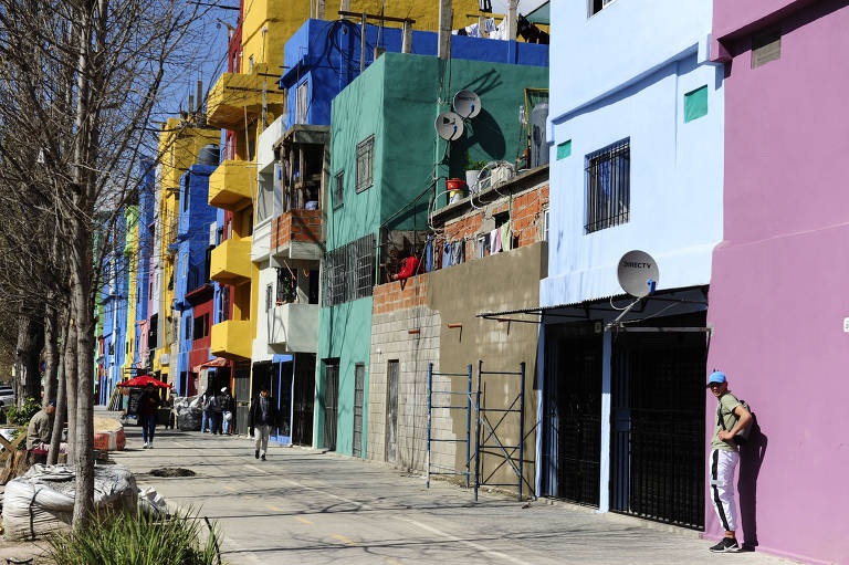 A imagem mostra uma rua com casas coloridas em tons de azul, verde, amarelo e rosa no que parece ser uma favela. As edificações têm diferentes estilos e algumas apresentam grafites. Há pessoas caminhando pela calçada e uma árvore sem folhas à esquerda. O céu está claro e ensolarado.