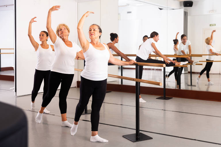 Mulheres de diferentes idades exercitando movimentos de balé na sala de treinamento.