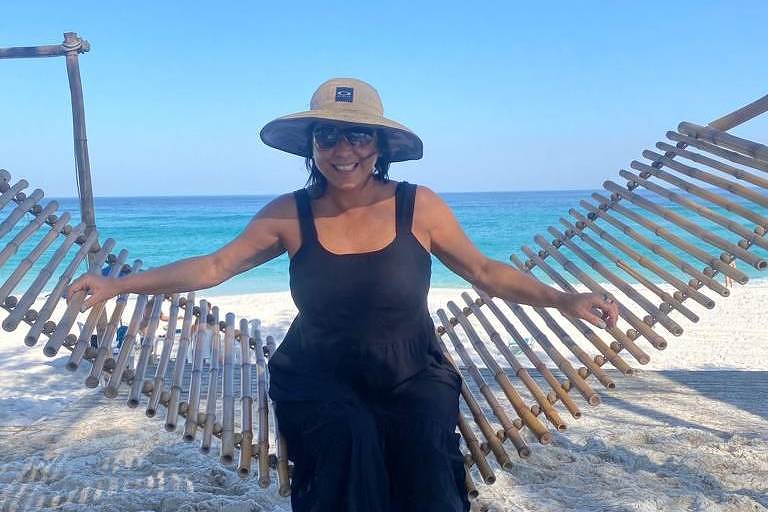 cristiane usa vestido preto, chapéu e óculos escuros. posa para a foto à frente de uma rede de madeira na praia. ao fundo, areia e o mar