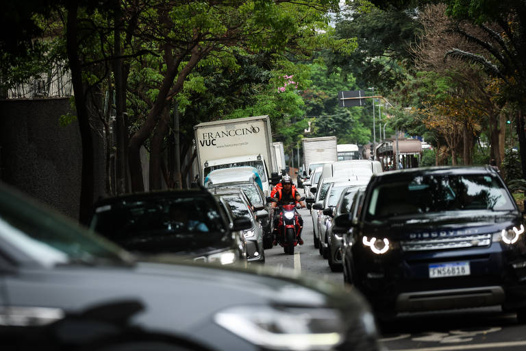 A imagem mostra uma rua movimentada com tráfego intenso. Vários carros estão parados ou em movimento， incluindo um caminhão e uma motocicleta. Árvores estão visíveis ao longo da calçada， proporcionando sombra. O ambiente parece urbano e a iluminação é natural， sugerindo um dia claro.