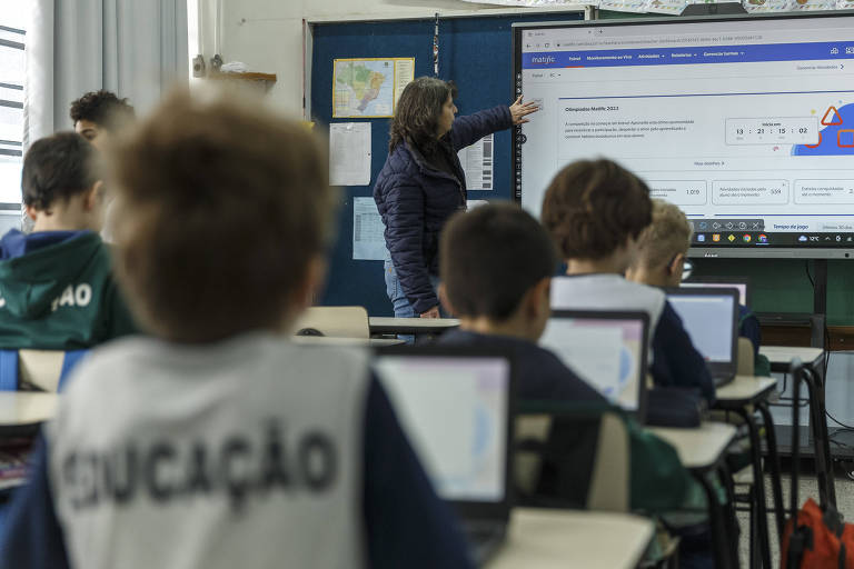 Imagem mostra alunos com notebooks sobre as carteiras durante uma aula. Ao fundo, a professora toca em uma lousa digital touchscreen
