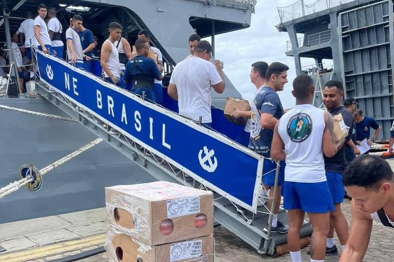 Imagem mostra homens carregando caixas para dentro do navio escola. É possível ver um banner azul com o dizer "NE Brasil" 