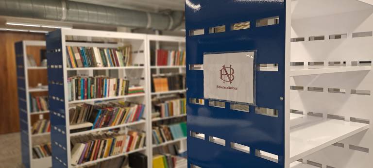 Imagem mostra estante azul e branca vazia, com uma placa da biblioteca nacional. Ao fundo, é possível ver estantes cheias de livros
