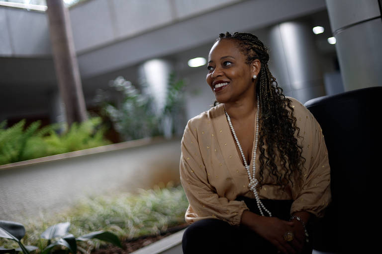 Imagem em primeiro plano mostra mulher negra posando para foto sorrindo. Ela está sentada e de perfil.