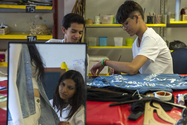 Dois meninos e uma menina estilizam jaquetas jeans em sala de artes. Menina aparece na foto através de reflexo em espelho