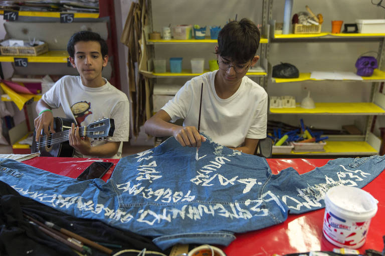 Dois meninos na foto, um toca violão e outro estiliza jaqueta jeans com escritos em tinta branca