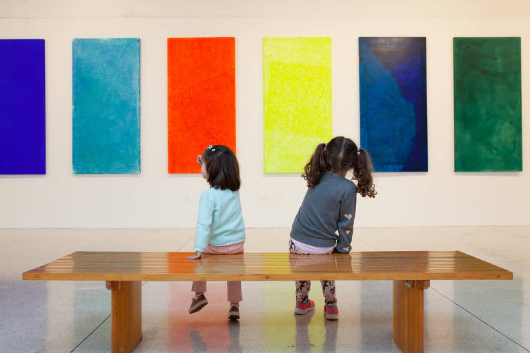 duas crianças sentadas em banco de madeira em sala clara observam seis telas com cores azul, vermelha amarela e verde