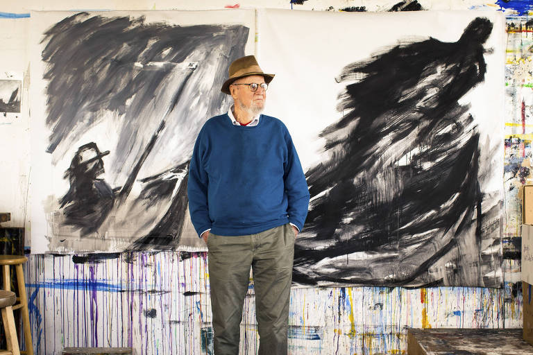 homem idoso branco com roupa azul e chapéu em frente a pintura em tons de preto