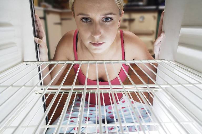 Mulher olhando para dentro de geladeira, cujas prateleiras estão vazias