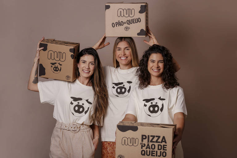 Três mulheres brancas com camisetas brancas seguram caixas de papelão da Nuu, com dizeres "pizza de paõ de queijo" "Nuu o pão de queijo"