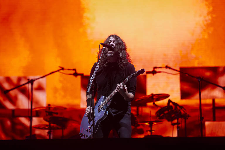 Homem, no centro da imagem, de cabelos longos, tocando uma guitarra azul e cantando. Atrás, parte da bateria e um fundo vermelho