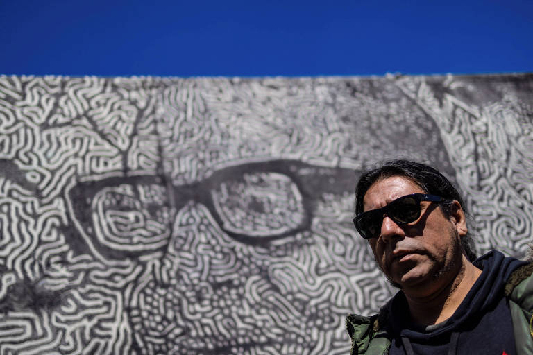 Fotografia colorida mostra homem de óculos escuros que passa por mural em que se vê desenhado o rosto de um homem com óculos quadrados grossos e um pedaço de céu azul  