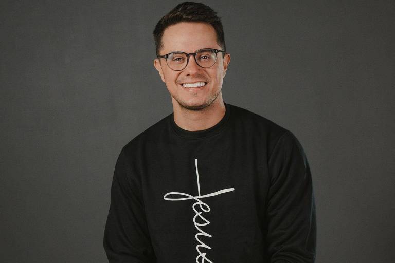 Homem branco de óculos sorrindo com blusa escrito 'jesus'