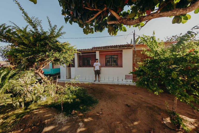 Homem vestindo bermuda preta e camiseta branca está diante do muro branco de uma casa térrea que tem na frente um pátio com árvores e chão de terra vermelha