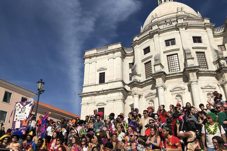 A imagem mostra um grande grupo de pessoas reunidas em frente a um edifício histórico em Lisboa, durante uma celebração de Carnaval. As pessoas estão vestidas com roupas coloridas e fantasias, algumas usando chapéus e acessórios divertidos. O céu está claro e azul, e o edifício ao fundo é de arquitetura clássica, com uma cúpula e janelas em destaque