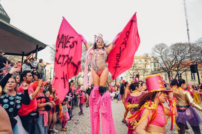 Desfile do bloco Colombina Clandestina. No centro da imagem, uma mulher andando com pernas de pau. Ela está com uma fantasia rosa e prateada e traz faixas com mensagens "Save Lives" e "Travesti"