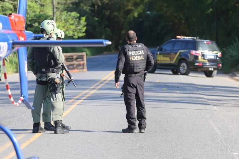 Foto mostra dois policiais federal de costas, uma viatura e parte de um helicóptero em uma rua 
