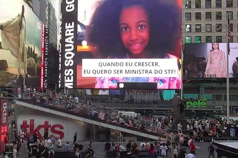 Um grande telão no meio de prédios e pessoas circulando mostra uma menina negra