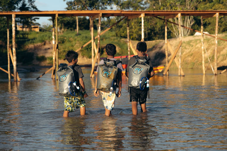 Três crianças, todos meninos, caminham em área de igarapé. Todo carregam nas costas do filtro de água da startup, em formado de mochila