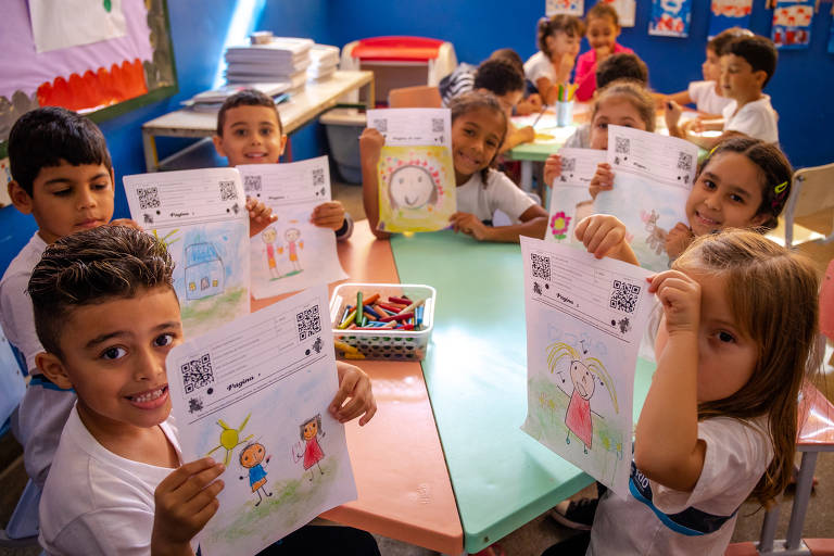 Sala de aula com mesas em formato circular acomoda dezenas de crianças que, sentadas, pintam e ilustram