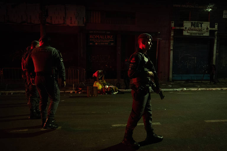 Policial militar portando arma longa, tingido de vermelho pela luz da viatura da PM, com dois policiais logo atrás dele e, nos fundos, uma rua suja, com comércios fechados e um morador de rua revirando o lixo