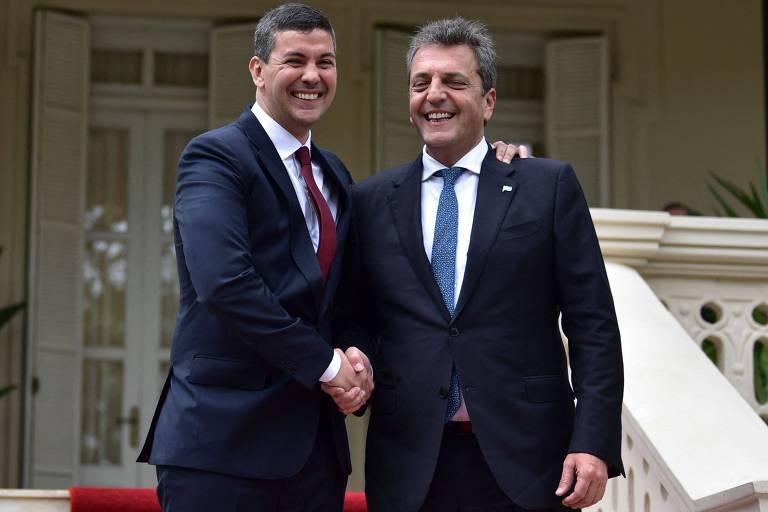 Dois homens de terno apertam a mão sorrindo em frente a palácio
