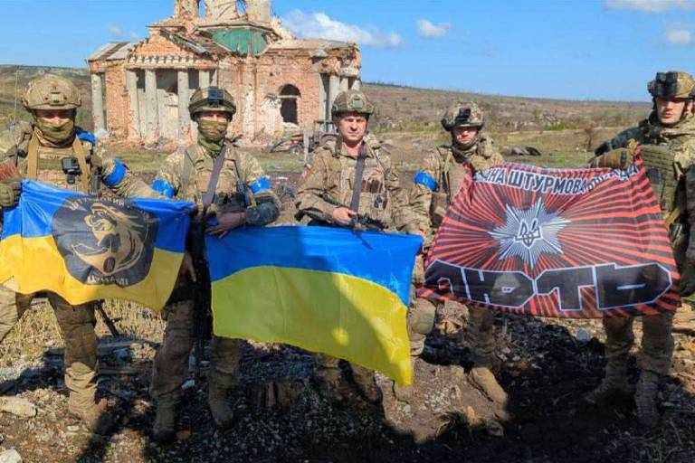 Militares ucranianos posam para foto com bandeiras em frente a um prédio destruído na vila de Klyschiivka, região de Donetsk