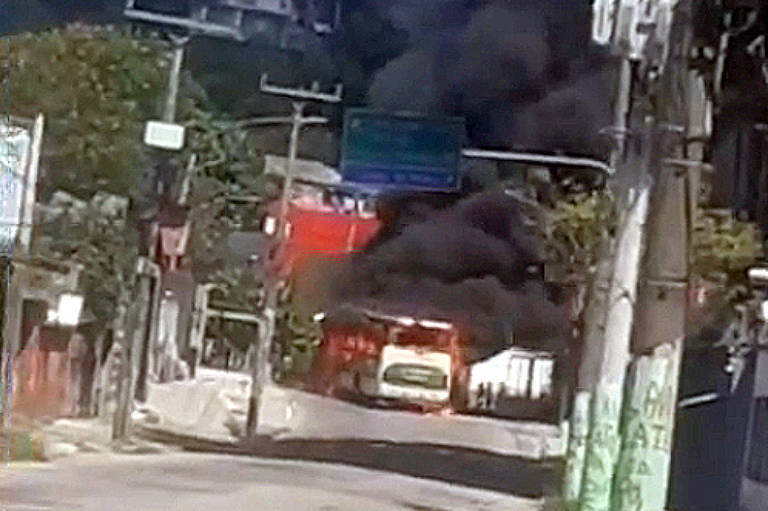 Rua asfaltada. Ao fundo, ônibus de pintura verde e branca pega fogo. Fumaça sai pelas janelas e pelo teto. A fumaça é preta e parece densa. O céu está azul