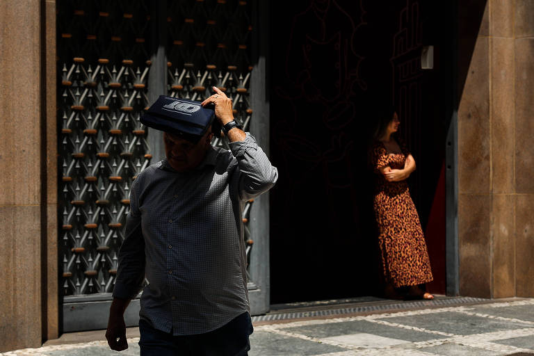 homem caminha e se protege do sol com pasta sobre a cabeça; e, ao fundo, mulher recostada se protege sob a sombra de um prédio