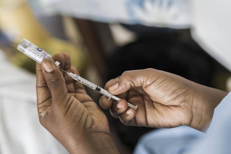 Imagem fechada em duas mãos puxando vacina de dentro de um vidro com uma seringa