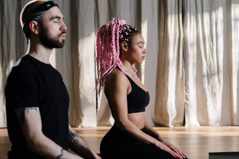 Um homem branco de camiseta preta e uma mulher negra, de tranças cor-de-rosa e top de academia preto, sentam em posição de meditação