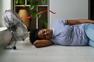Overheated African woman sleeping on floor with electric fan on, suffering from heat at home