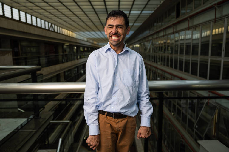 Um homem usando camisa de manga comprida azul clara e calça caqui, sorri largamente. Ele tem cabelos pretos e barba por fazer. Atrás dele, várias janelas de vidro