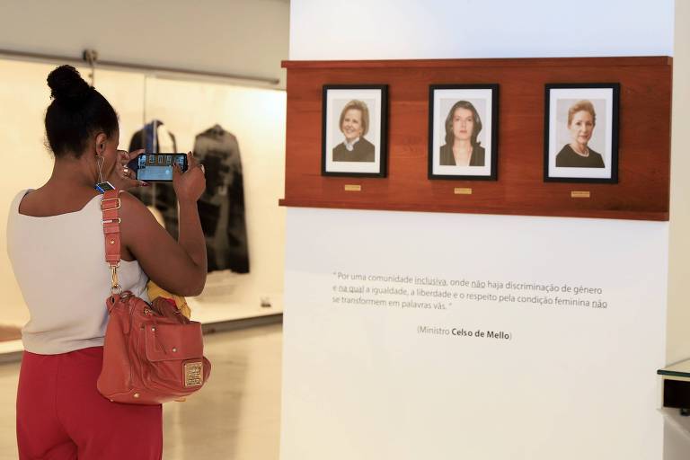 Mulher negra tira fotografia de painel branco com retratos das três ministras