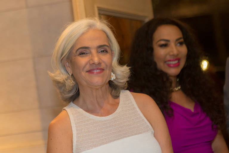 A imagem mostra duas mulheres em um ambiente interno. A mulher à esquerda tem cabelo grisalho e usa uma blusa branca com detalhes em renda. Ela sorri para a câmera. A mulher à direita tem cabelo cacheado e usa um vestido rosa， também sorrindo. O fundo é desfocado， sugerindo um evento social