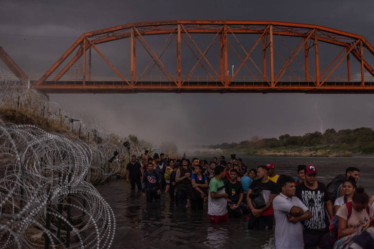Raios iluminam e poeira sopra ao fundo de uma tempestade iminente, enquanto centenas de migrantes se alinham sob a ponte de trem Puente Negro Ferrocarril, aguardando para se renderem às autoridades após atravessarem o rio Grande do México para os Estados Unidos em Eagle Pass, Texas, EUA