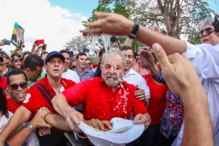 Lula, de camisa vermelha, cercado por apoiadores em local a céu aberto. Alguém joga água à frente