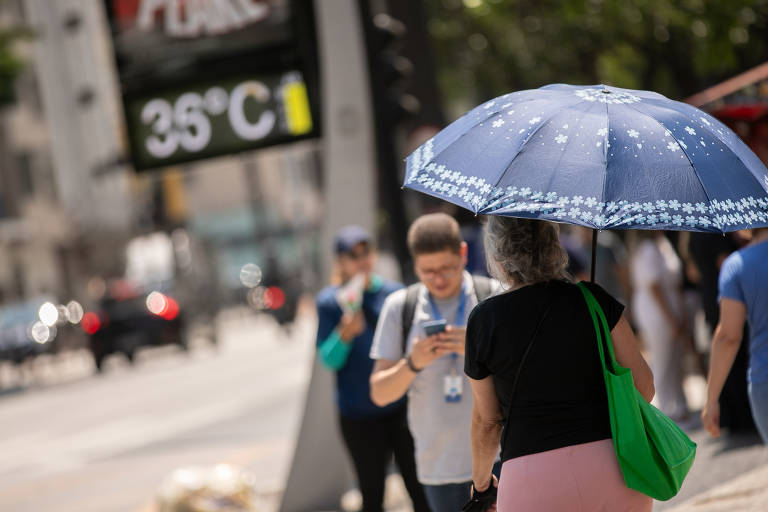 Pedestres caminham em rua com carros ao fundo; em primeiro plano, uma mulher de costas com um guarda-chuva para se proteger do sol. Mais ao fundo, termômetro marcando 35 ou 36°C (não é possível ler com definição)