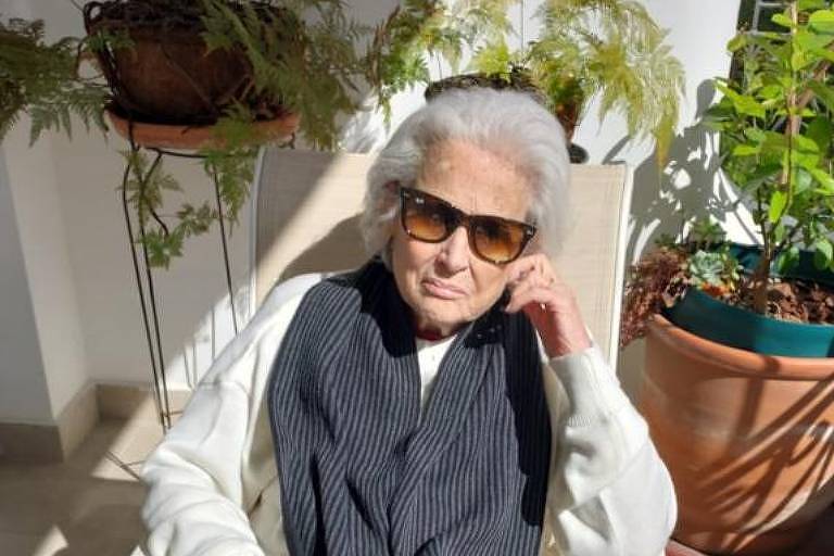Mulher idosa, de cabelos brancos e óculos de sol, sentada em uma cadeira em uma varanda
